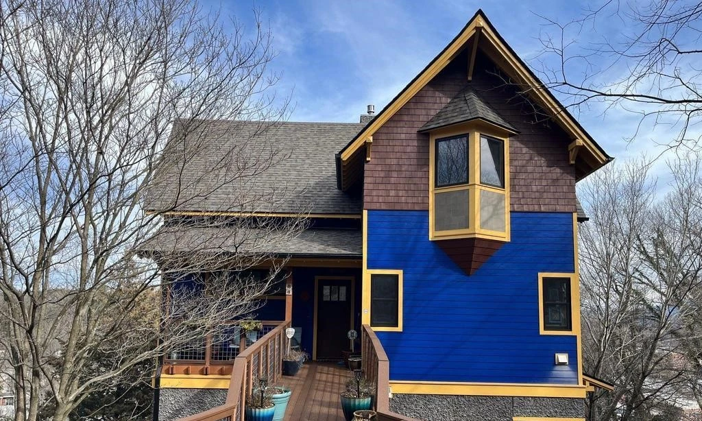  A vibrant modern home painted in blue and dark brown with contrasting shingle siding. The house has a unique design with a steeply pitched roof, large windows, and a wooden porch. Surrounded by leafless trees, the home stands out in a serene neighborhood setting.