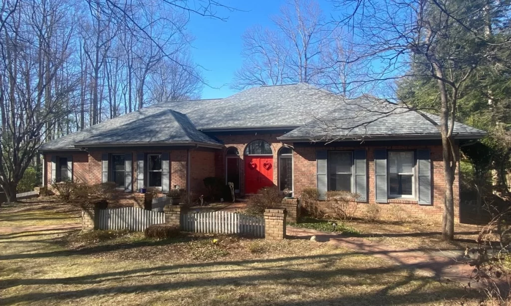 A traditional ranch-style home featuring a red brick exterior and a gray shingle roof. The house has a prominent red front door decorated with heart-shaped wreaths, and is surrounded by a small picket fence and bare trees, giving it a welcoming, suburban look.